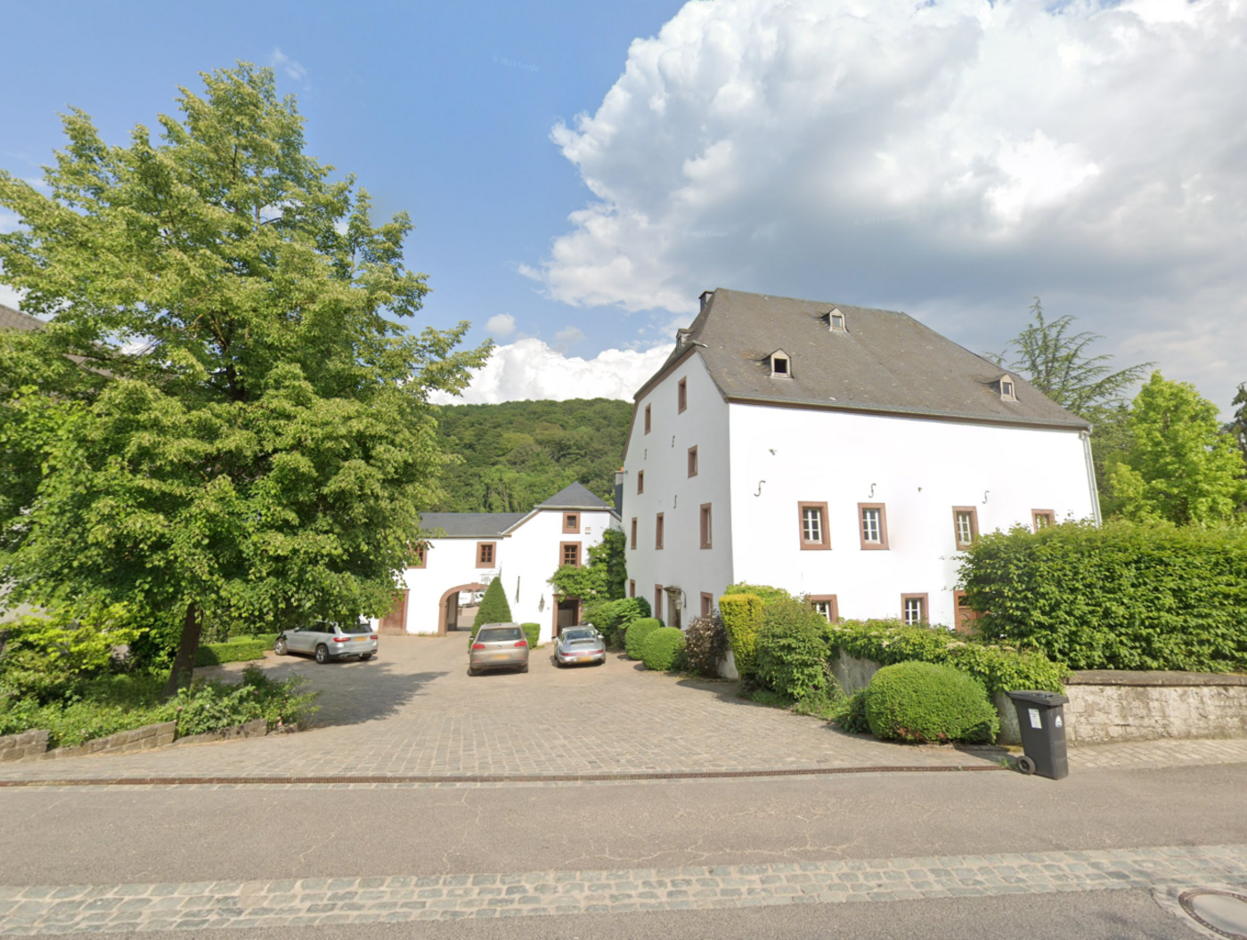 Photo of a white manor house with a paved courtyard and parked cars, surrounded by trees