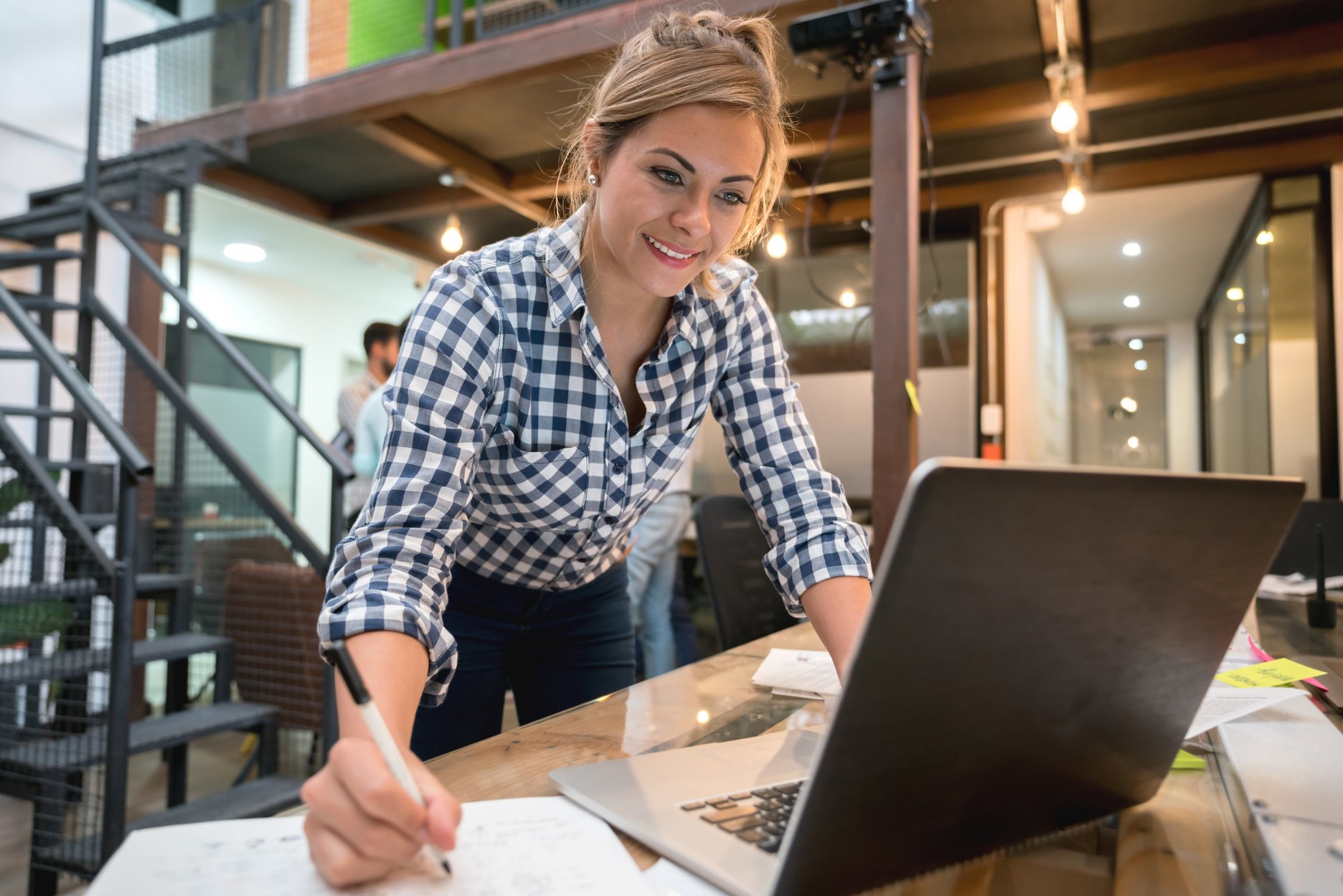 A woman leaning over a desk, writing in a notebook while looking at a laptop in a modern office setting.
