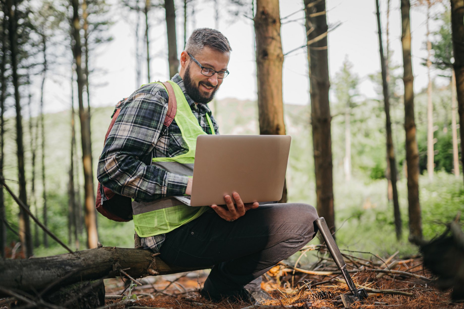 Ein Mann kniet in einem Wald, trägt eine Warnweste und arbeitet an einem Laptop.